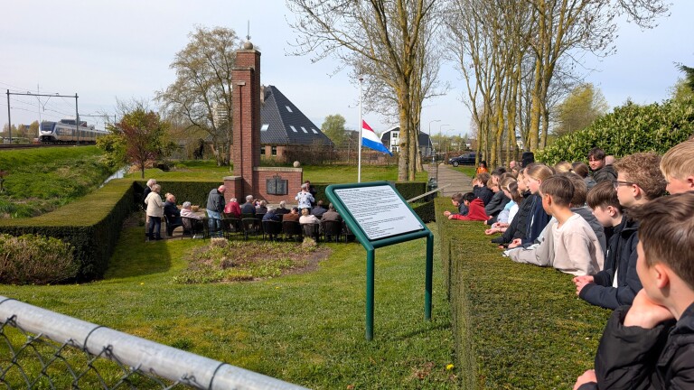 Groep mensen bij een gedenkteken naast een spoorlijn, met een Nederlandse vlag halfstok en kinderen kijkend op de achtergrond.