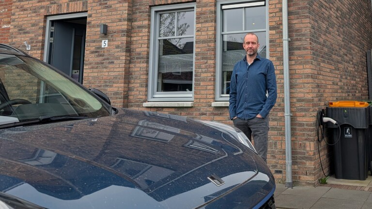 Man in een blauwe blouse staat voor een huis met bakstenen muren naast een geparkeerde auto en een afvalcontainer.