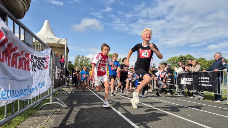 Kinderen rennen op een racebaan tijdens een sportevenement, omringd door toeschouwers en hekken met banners.