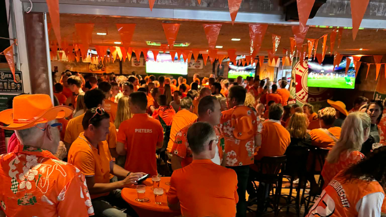 Grote groep mensen in een café, gekleed in oranje, kijkt naar een voetbalwedstrijd op meerdere schermen met oranje vlaggetjes erboven.
