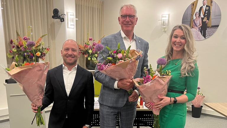 Drie mensen met blije gezichten houden boeketten vast in een kamer, met een foto van koninklijk paar op de achtergrond.