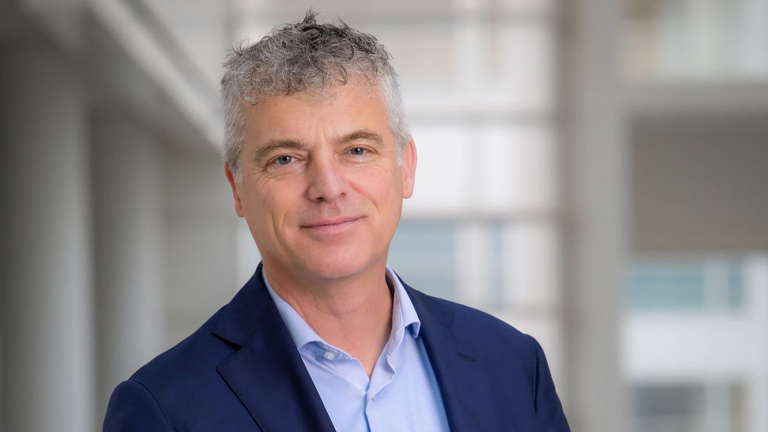 Man met grijs haar en blazer, glimlachend in een lichte, moderne ruimte.