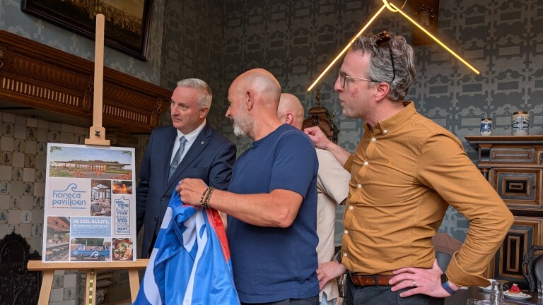 Groep mannen kijkt naar een presentatiebord met afbeeldingen en tekst over een horecapaviljoen, in een sierlijke kamer.