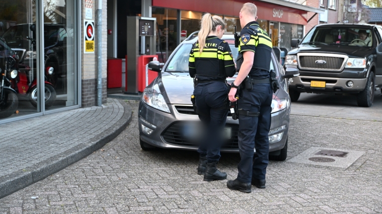 Twee politieagenten in gesprek bij een geparkeerde auto voor een winkel met geblurde kentekenplaat.