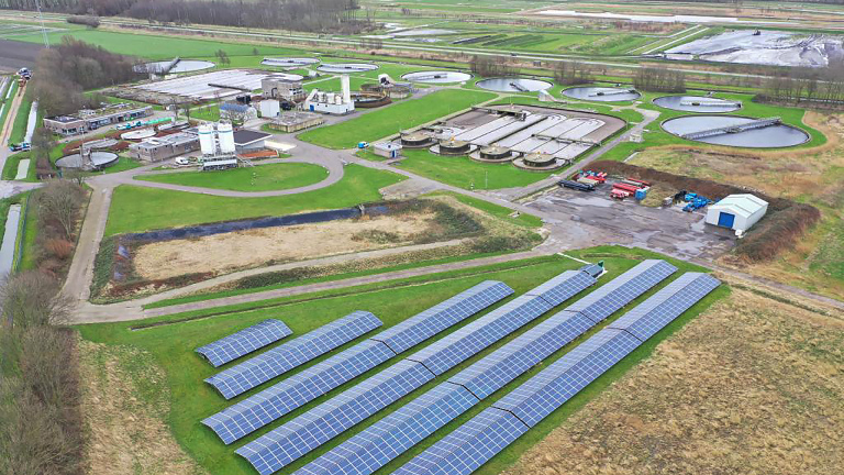 Zonnepanelen op een veld naast een waterzuiveringsinstallatie omringd door groene landschappen.
