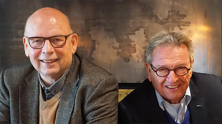 Twee oudere mannen lachen naar de camera, zittend voor een muur met een wereldkaart.