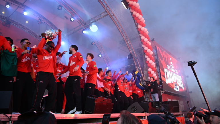 Een team viert feest op een podium met een trofee, in rood gekleed met het woord "CHAMPIONS" op hun shirts.