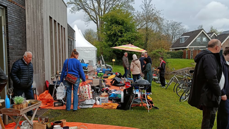 Mensen bezoeken een rommelmarkt buiten, met tafels vol spullen, onder een kleurrijke parasol.