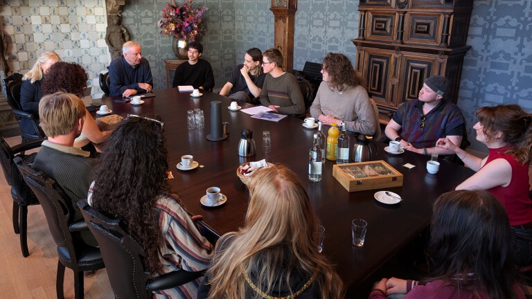 Mensen zitten rond een grote tafel in een vergaderruimte met koffie en drankjes, terwijl een oudere man spreekt.