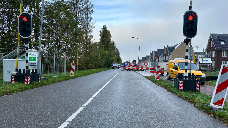 Twee verkeerslichten op rood bij een wegafsluiting met borden en wegafzettingen, een gele auto geparkeerd aan de rechterkant, en huizen aan de rechterzijde van de weg.