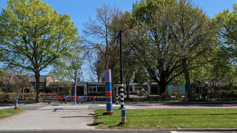 Kleurrijke palen en zebrapaden leiden naar een schoolgebouw omringd door bomen met een heldere blauwe lucht erboven.