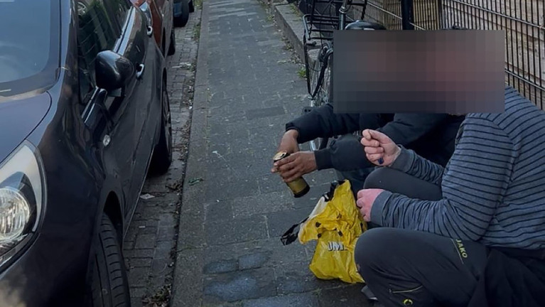 Twee mensen zitten op een smalle stoep naast een geparkeerde auto. De een houdt een blikje drank vast en er ligt een gele plastic tas op de grond. Hun gezichten zijn gepixeleerd.