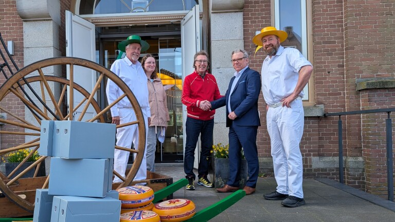 Een groep mensen, sommigen in witte kleding met hoeden, staan buiten bij een gebouw naast kazen en kratten. Twee mannen schudden elkaar de hand.