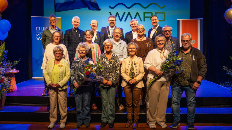 Groep mensen poseert op een podium, sommigen dragen onderscheidingen; bloemen en banners zijn zichtbaar op de achtergrond.