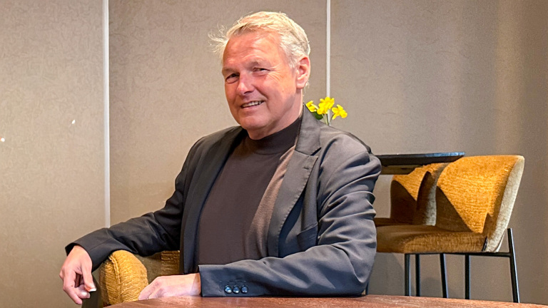 Een man in een donkere trui en jasje zit aan een tafel, met gele bloemen op de achtergrond.