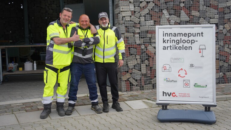Drie mannen in fluorescerende werkkleding poseren lachend naast een bord met de tekst "innamepunt kringloop-artikelen".
