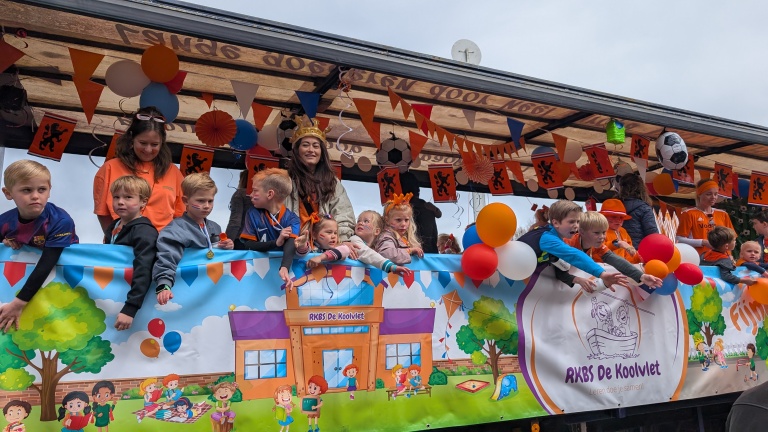 Een versierde wagen met kinderen en volwassenen in oranje kleding, omringd door ballonnen en oranje vlaggetjes.