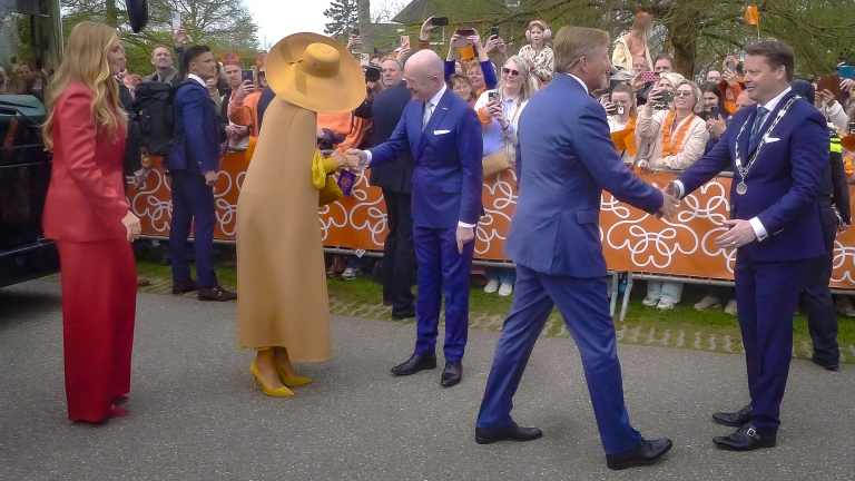 Mensen in chique kleding schudden handen op een openbare bijeenkomst met veel omstanders en oranje decoratie op de achtergrond.