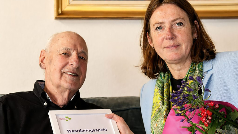 Een oudere man en een vrouw houden een certificaat met de tekst "Waarderingsspeld" vast; de vrouw heeft een boeket bloemen.