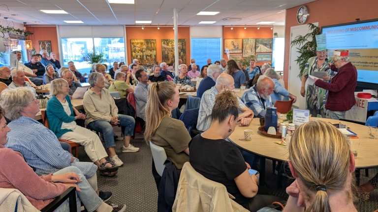 Een groep mensen bij een presentatie in een zaal, met aandacht voor een scherm waarop "Misleiding of Miscommunicatie?" staat.