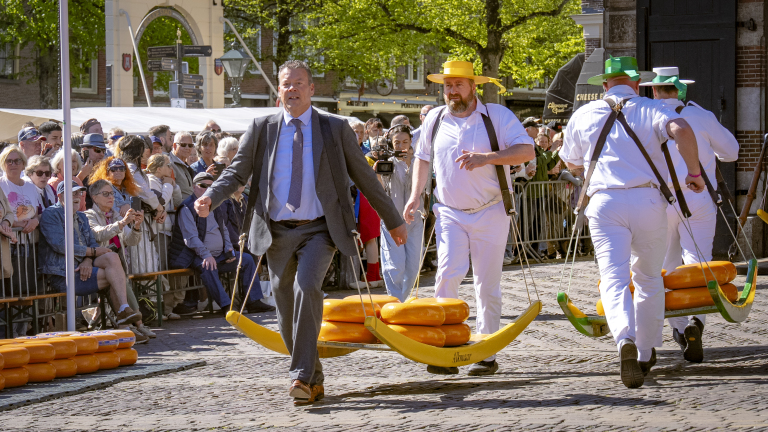 Mannen dragen kaas op traditionele dragers tijdens een festival, omringd door toeschouwers op een zonnige dag.