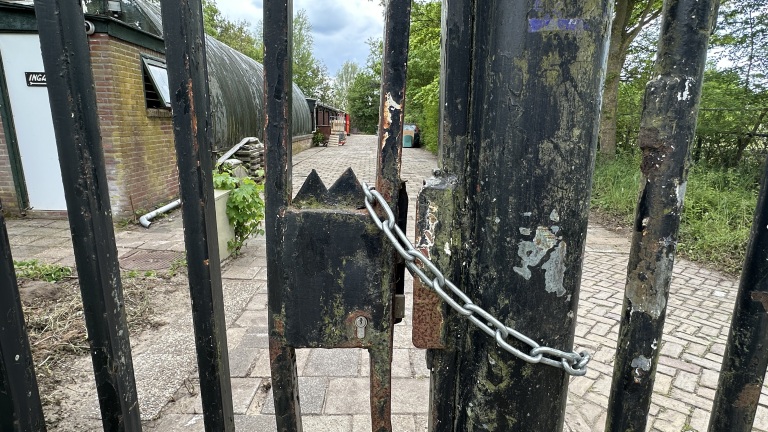 Verroest hek met ketting en slot, zicht op gebouw met bordje "Ingang" en een verhard pad tussen bomen.