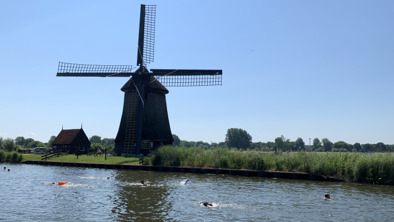 Windmolen naast een kanaal met zwemmers, omringd door groen landschap onder een blauwe hemel.