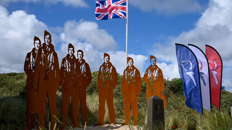 Zeven metalen silhouetten van piloten staan onder een Britse vlag in een grasveld, met vlaggen aan de zijkant.