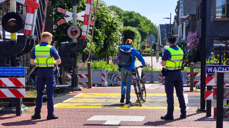 Twee handhavers bewaken een spoorwegovergang terwijl een fietser met fiets passeert.