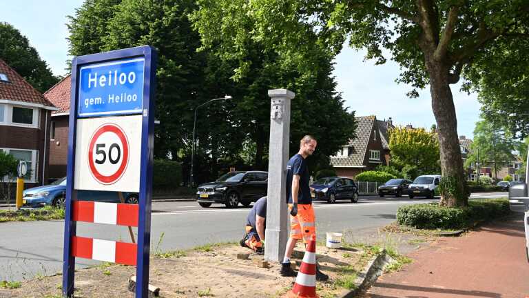 Verkeersbord met "Heiloo" en snelheidslimiet 50 km/u, naast een man die werkt aan een paal op een zonnige straat.