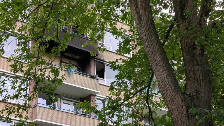 Verbrande verdieping van een gebouw, met gebarsten ramen en donkere schroeiplekken, omgeven door groene bomen.