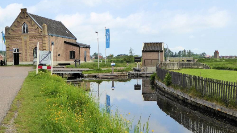 Bakstenen gebouw naast een kanaal met houten brug en grasvelden, onder een blauwe lucht met enkele bewolking.