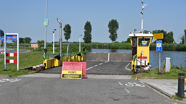 Pont is gestremd met waarschuwingsbord en verkeersborden, gelegen aan een rustige waterkant met bomen en natuur op de achtergrond.