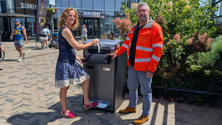 Een vrouw en een man bij een grijze afvalcontainer; de vrouw deponeert afval, de man draagt een feloranje jas.