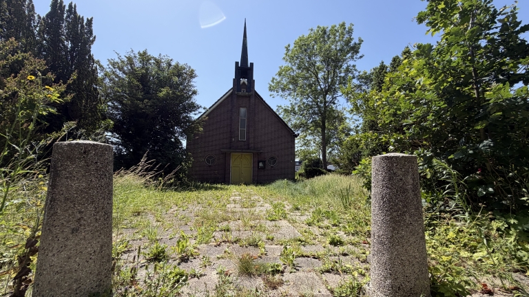Kleine bakstenen kerk met torenspits, omgeven door bomen en gras, gezien vanaf een pad met betonnen palen.