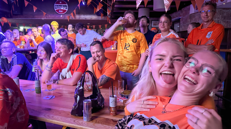 Twee lachende vrouwen in een drukke, feestelijke ruimte, omringd door mensen in oranje shirts.