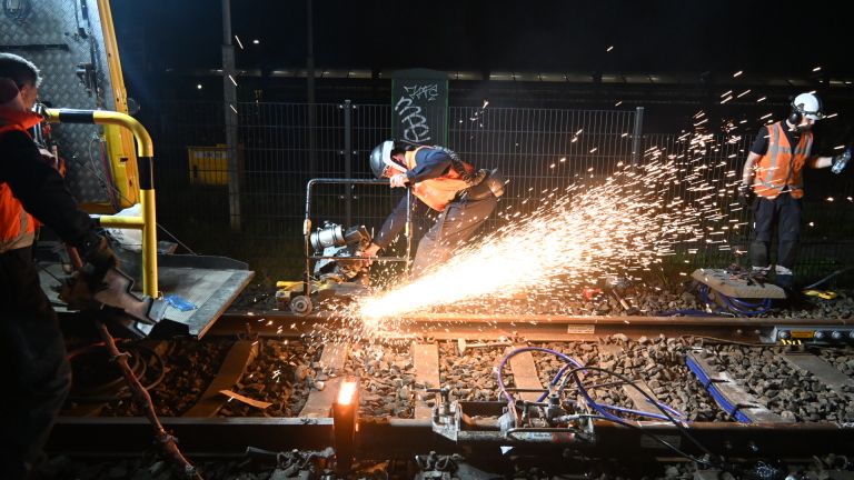Werkers in veiligheidsvesten snijden 's nachts met een slijpmachine door een spoorrail, omringd door vonken.