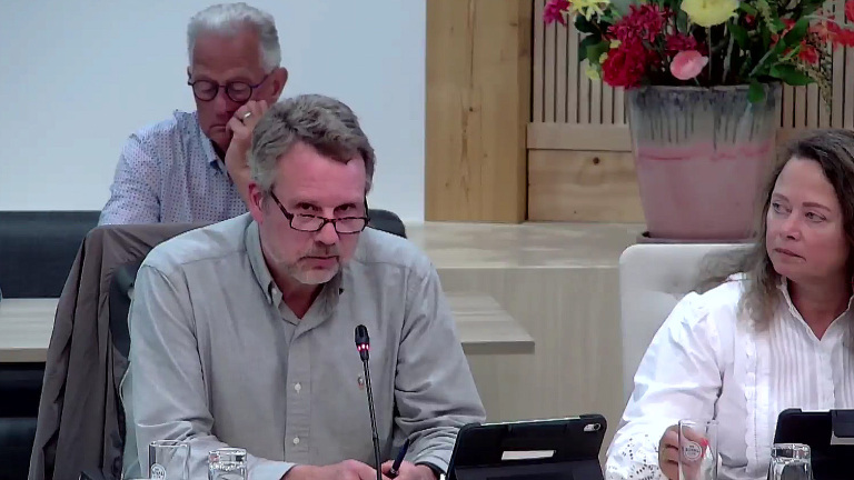 Een man spreekt in een microfoon tijdens een vergadering, met een vrouw naast hem en een vaas met bloemen op de achtergrond.