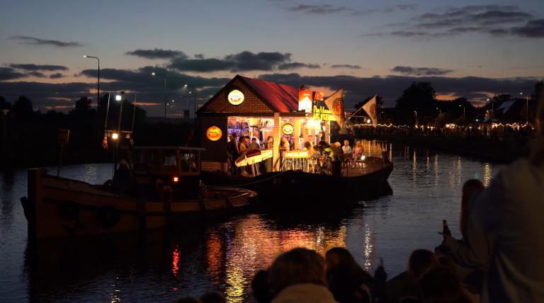 Een verlicht drijvend café op een kanaal tijdens de avondschemering, met mensen die bijeenkomen onder Amstel Bier-logo's, terwijl toeschouwers langs de oever zitten.
