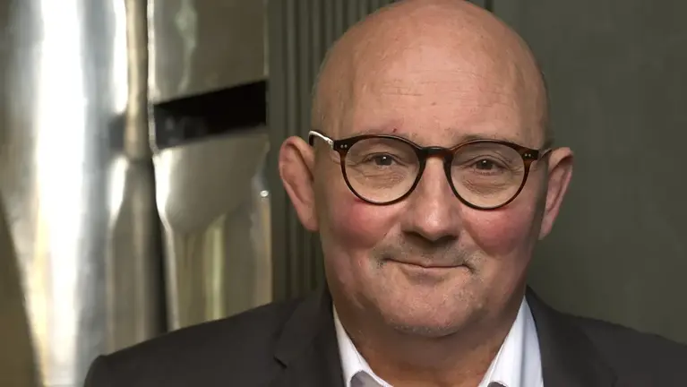 Man met bril, kaal hoofd en een glimlach, gekleed in een donker pak en wit overhemd, met een glanzende achtergrond.