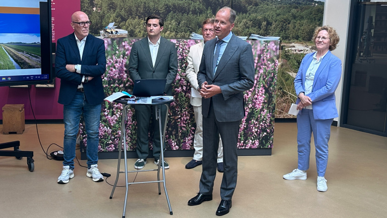 Een groep mensen staat bij elkaar in een ruimte met een achtergrond van bomen en bloemen, terwijl één man spreekt.