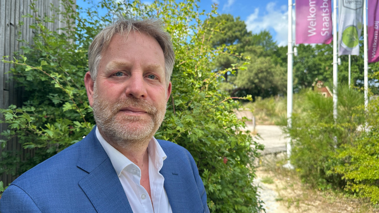 Man in een blauw pak staat buiten voor een groene struik, met vlaggen en tekst "Welkom Staatsbos" op de achtergrond.