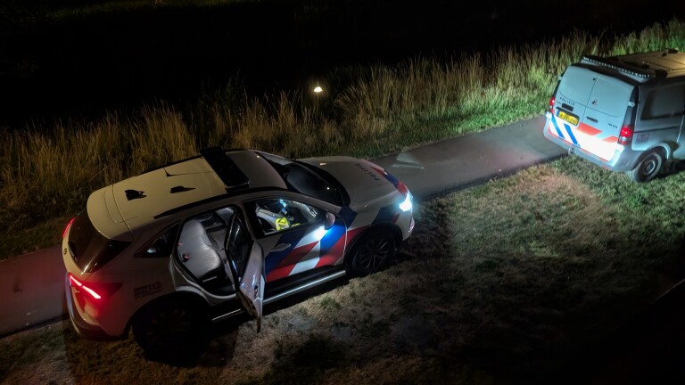 Twee politievoertuigen staan geparkeerd in het donker op een smal pad naast gras en hoge begroeiing.