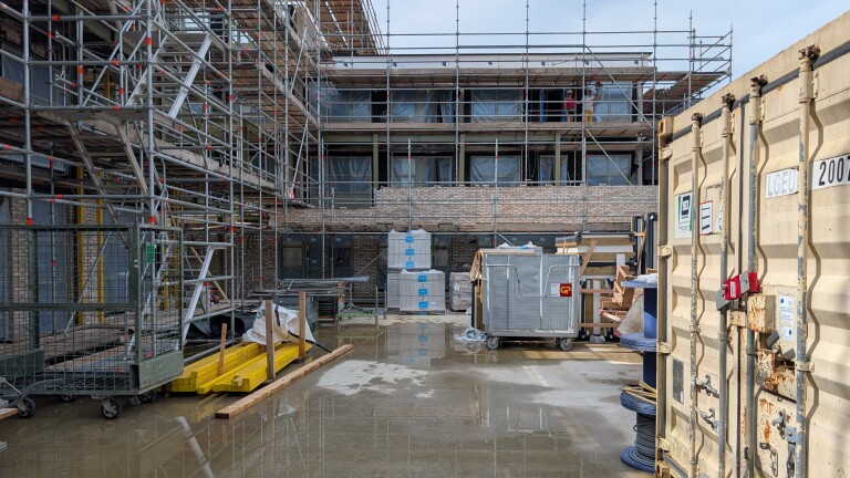 Bouwplaats met een gebouw in de steigers, bouwmaterialen en containers op de grond bij wisselend weer.