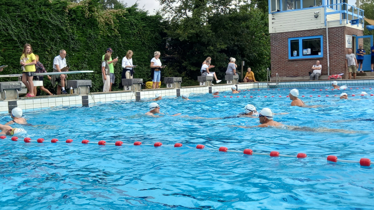 Zwemmers met witte badmutsen in een buitenbad, terwijl toeschouwers langs de rand toekijken bij startblokken