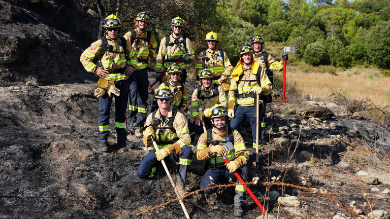 Groep brandweerlieden in beschermende kleding poseert met gereedschap op zwartgeblakerde grond na een natuurbrand