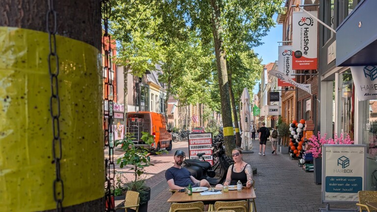 Twee mensen zitten aan een terrastafel in een winkelstraat met bomen, fietsen en winkelborden, waaronder HatShoe