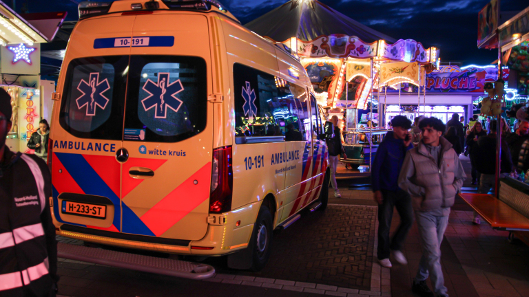Een ambulance staat geparkeerd op een verlichte kermis in de avond, omringd door mensen en attracties.