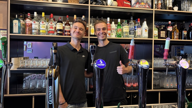 Twee barmedewerkers glimlachen achter de bar met bierpompen, met flessen sterke drank en glazen op de achtergrond