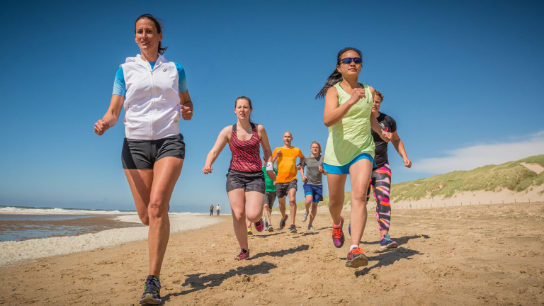Groep hardlopers rent over het strand langs de zee en duinen onder een heldere blauwe lucht